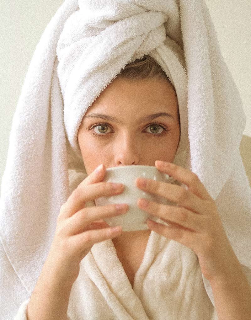 Woman in white towel robe drinking from a cup.