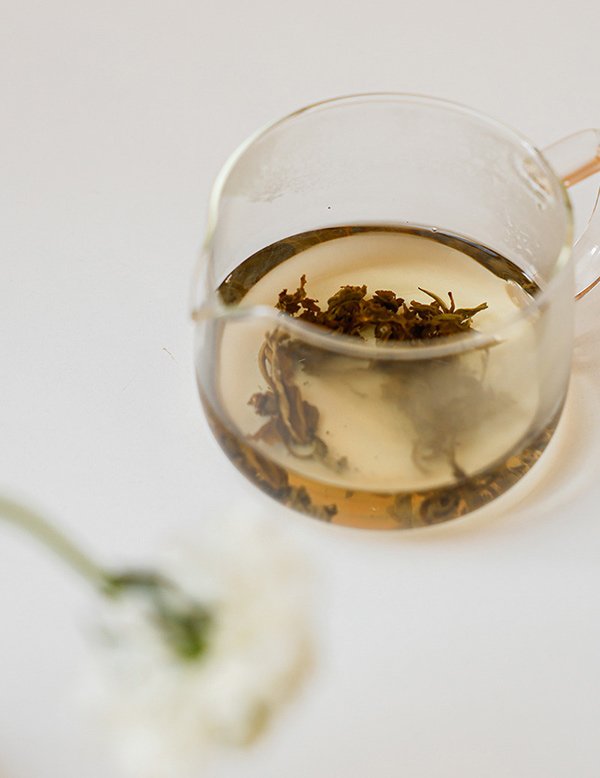 Glass teapot with steeping tea leaves on a white surface.
