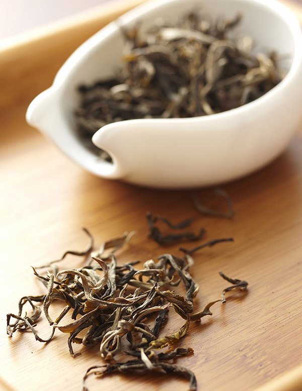 Loose dried tea leaves spilling from a white ceramic bowl onto a wooden surface.