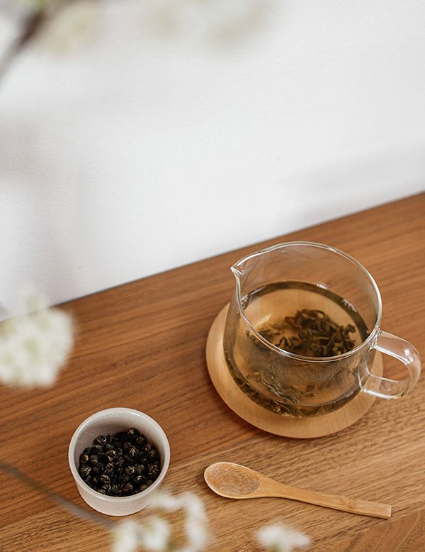 A glass teapot with brewed tea on a wooden coaster beside loose tea leaves.